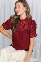 Woman wearing a red lace blouse sitting against a white curtain.