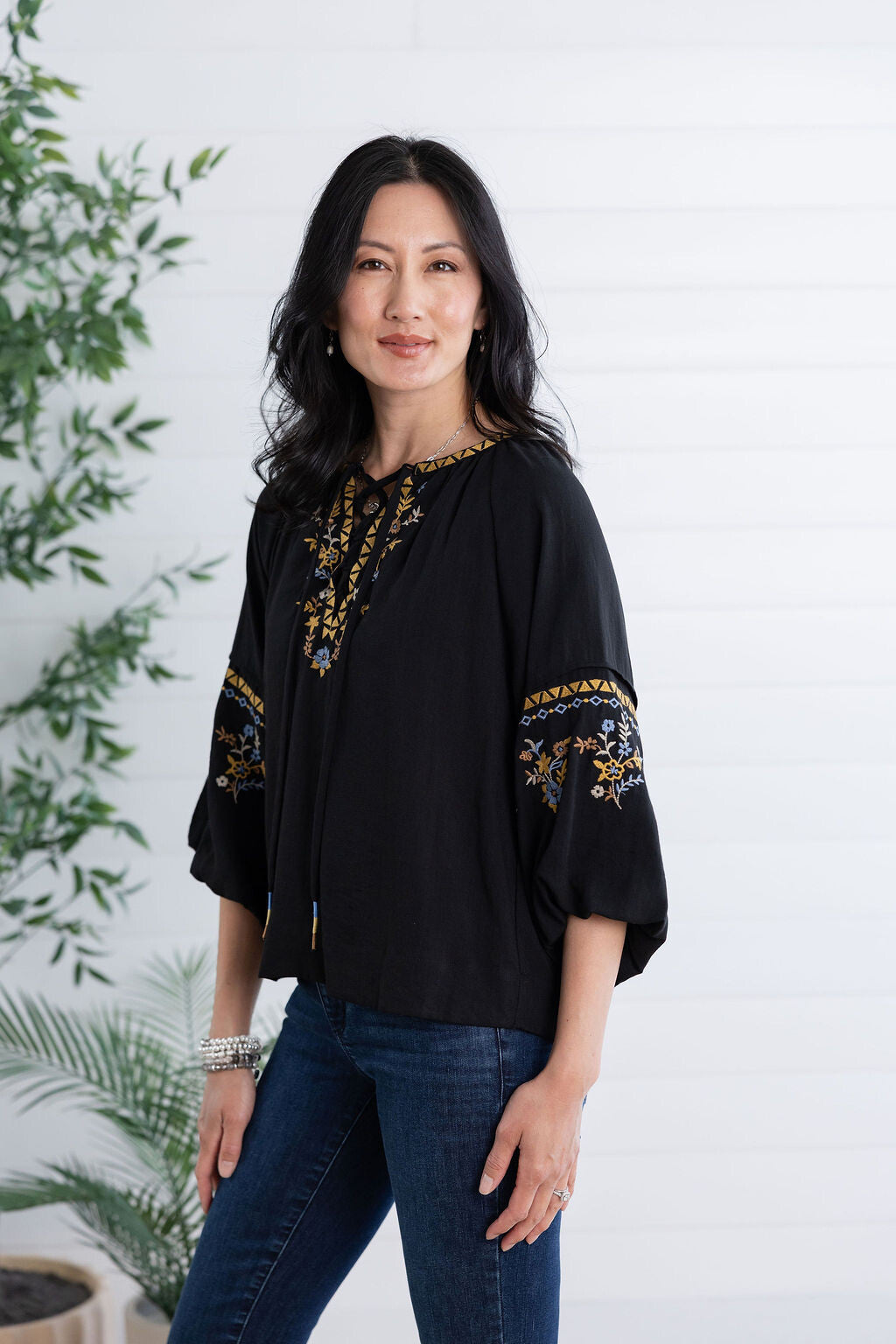 A woman modeling a black blouse with gold and blue floral embroidery on the neckline and sleeves, paired with dark blue jeans, standing against a white paneled wall with green plants on the side.
