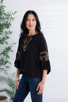 A woman modeling a black blouse with gold and blue floral embroidery on the neckline and sleeves, paired with dark blue jeans, standing against a white paneled wall with green plants on the side.