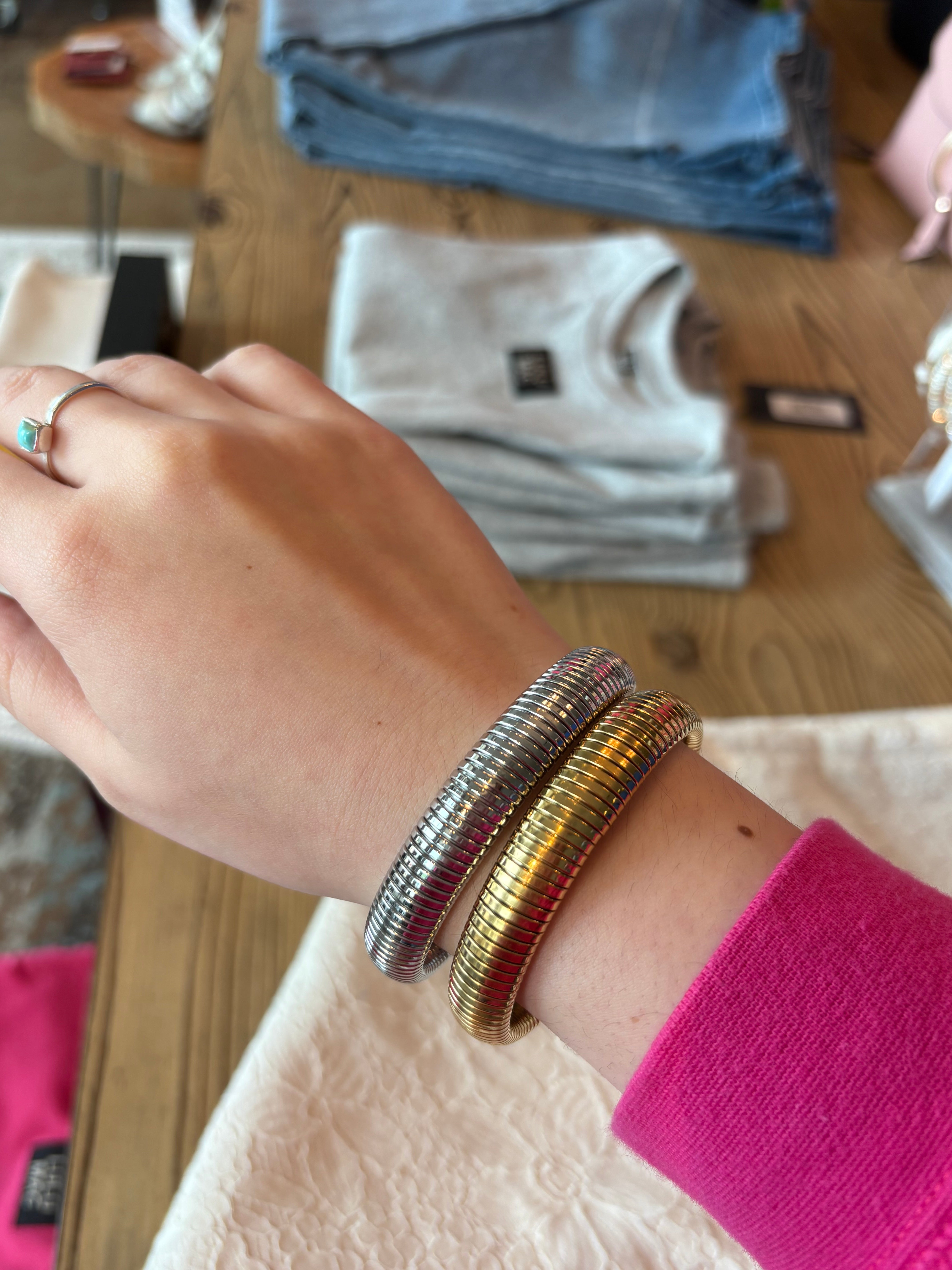A close-up of a person's wrist wearing two slim metallic bangles, one silver and one gold, with a bright pink sleeve visible. The background shows a wooden table with folded clothing in a store setting.