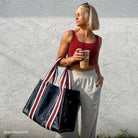 Woman holding a striped bag and a drink against a white wall