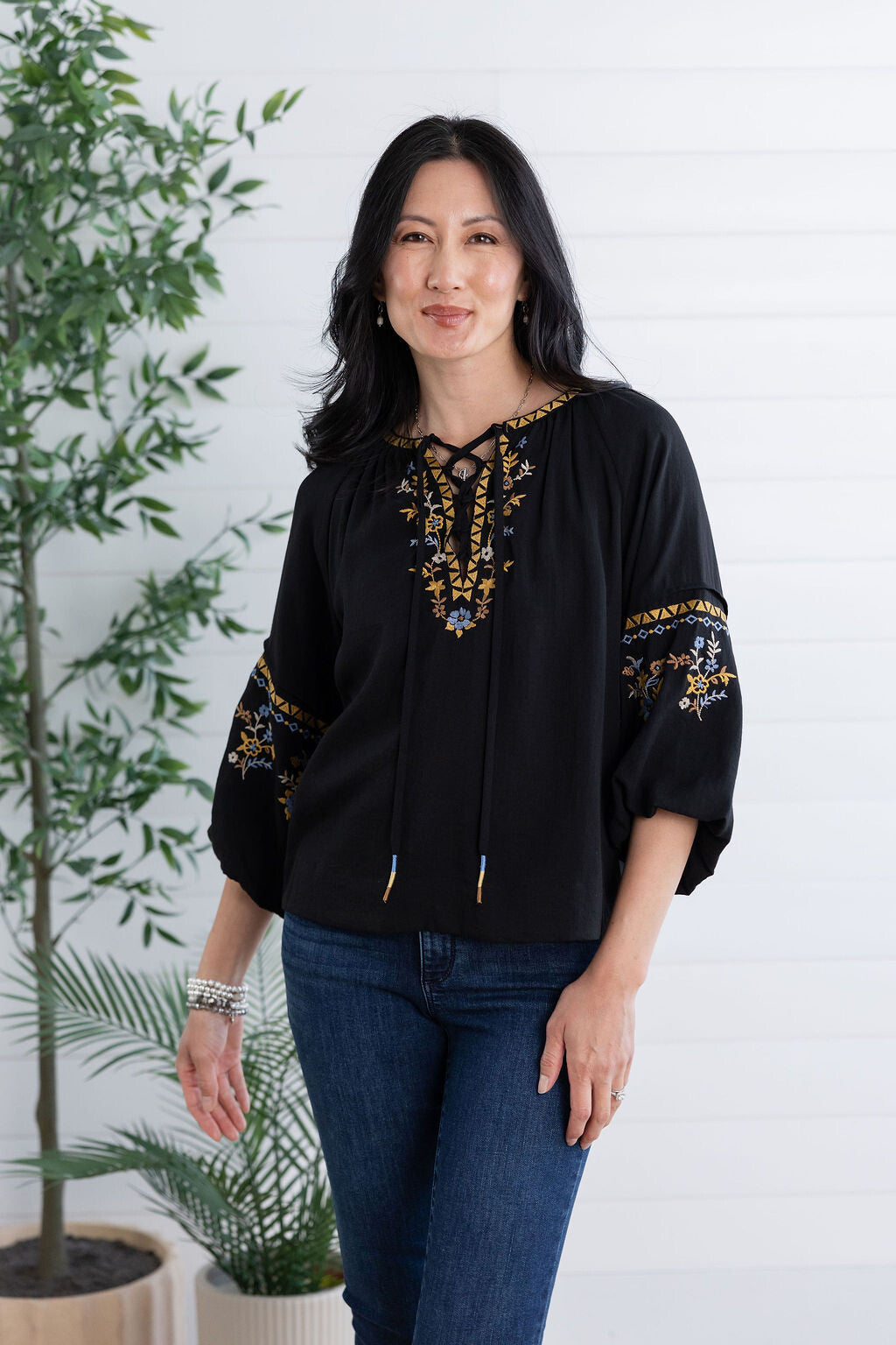 A woman modeling a black embroidered blouse with gold and blue detailing, paired with blue jeans, standing against a white paneled wall with potted plants.