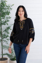 A woman modeling a black embroidered blouse with gold and blue detailing, paired with blue jeans, standing against a white paneled wall with potted plants.