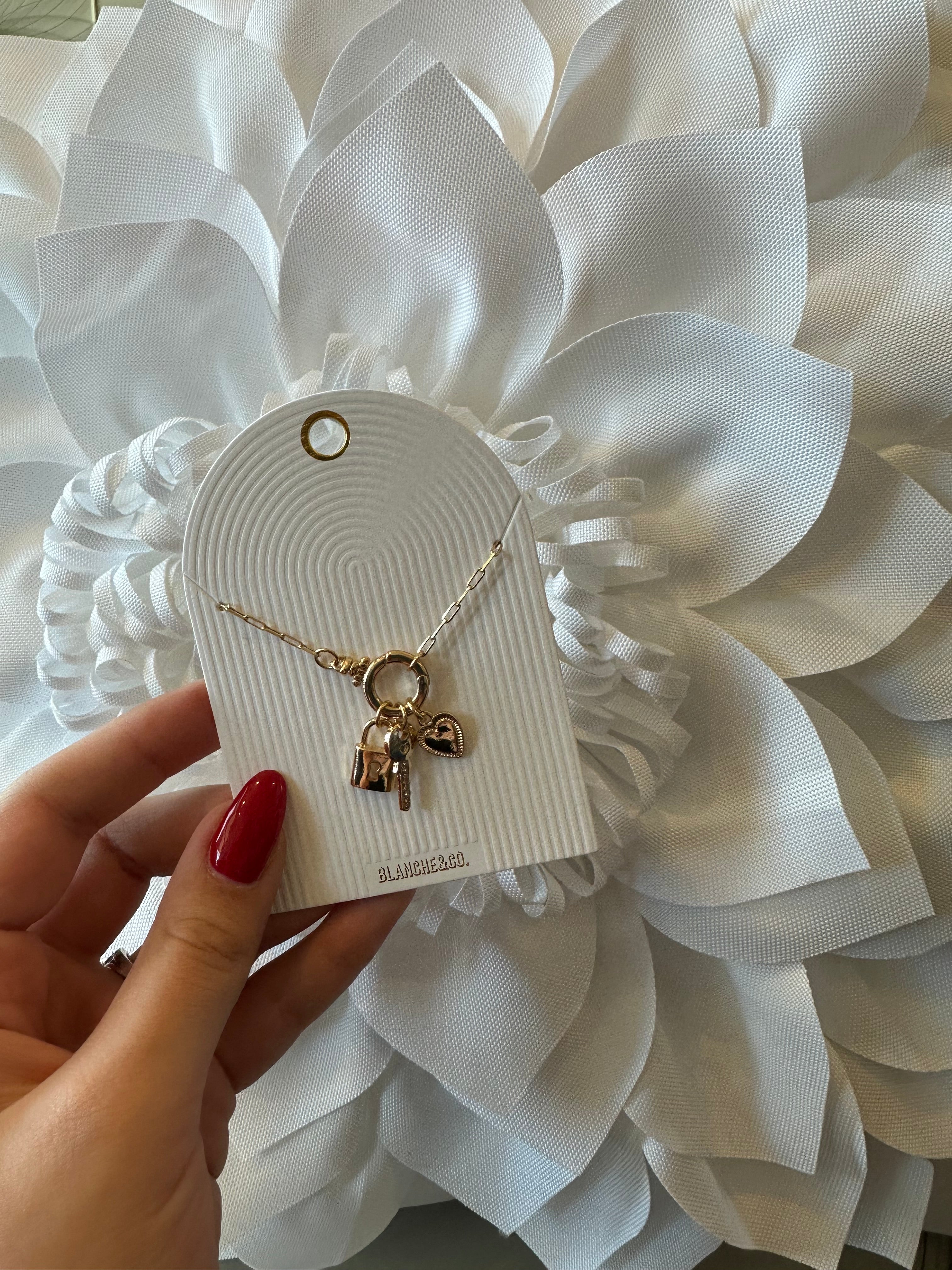 Gold necklace with heart and lock charms on a white card, held against a decorative white flower background.