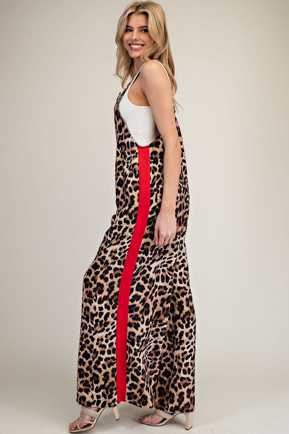 A woman modeling a sleeveless leopard-print maxi dress with a bold red side stripe, worn over a white tank top, photographed against a plain beige studio background.