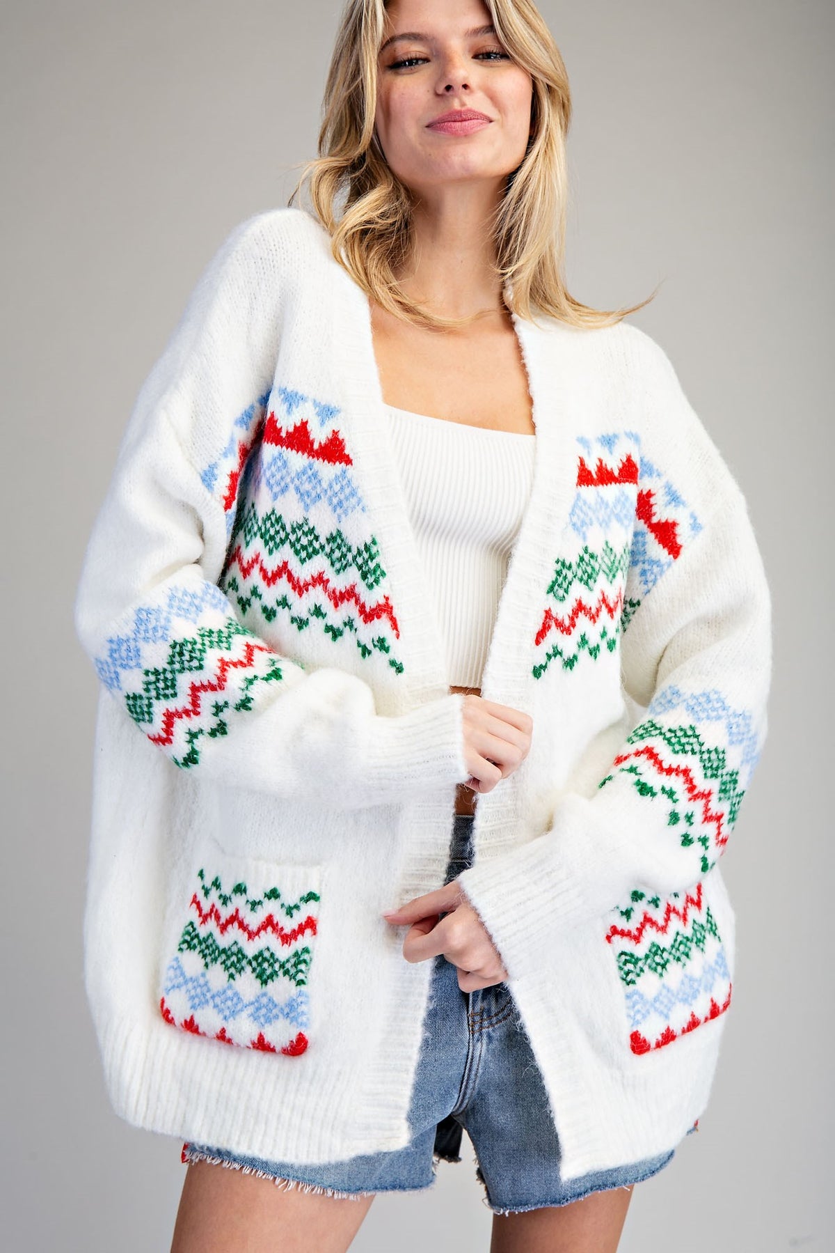 A woman wearing a white open-front knit cardigan with colorful Fair Isle geometric patterns in red, green, and light blue, styled with a white ribbed tank top and denim shorts against a neutral gray background.
