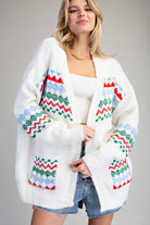 A woman wearing a white open-front knit cardigan with colorful Fair Isle geometric patterns in red, green, and light blue, styled with a white ribbed tank top and denim shorts against a neutral gray background.
