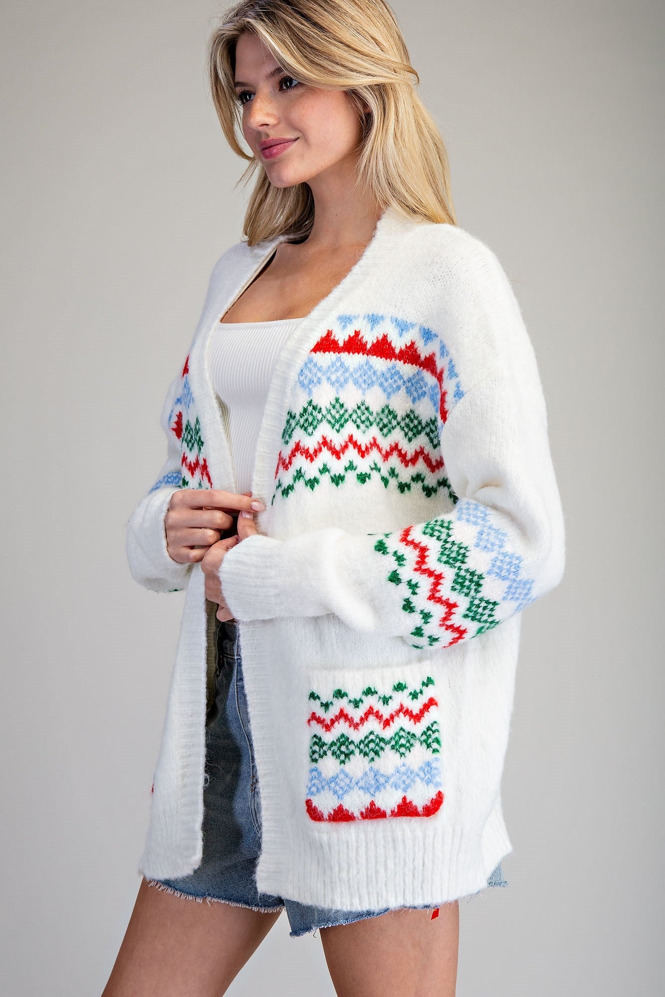 A woman wearing a white open-front knit cardigan with colorful Fair Isle style patterns in red, green, and light blue, styled over a white top and denim shorts against a plain gray studio background.