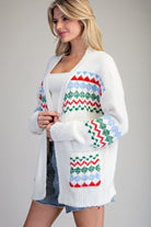 A woman wearing a white open-front knit cardigan with colorful Fair Isle style patterns in red, green, and light blue, styled over a white top and denim shorts against a plain gray studio background.
