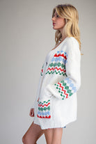 A woman wearing a white fuzzy knit cardigan with colorful Fair Isle patterns in red, green, and light blue, shown from a side profile against a plain light gray studio background.