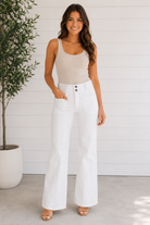 Woman wearing a beige tank top and white wide-leg pants standing against a white wall.