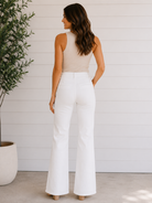 Woman wearing a beige top and white pants standing against a white wall with a plant to the left.