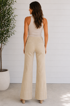 Woman wearing a beige sleeveless top and wide-leg pants standing in a minimalistic room with a plant.
