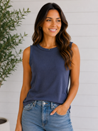 Woman wearing a navy sleeveless top and blue jeans standing against a white wall with a plant.