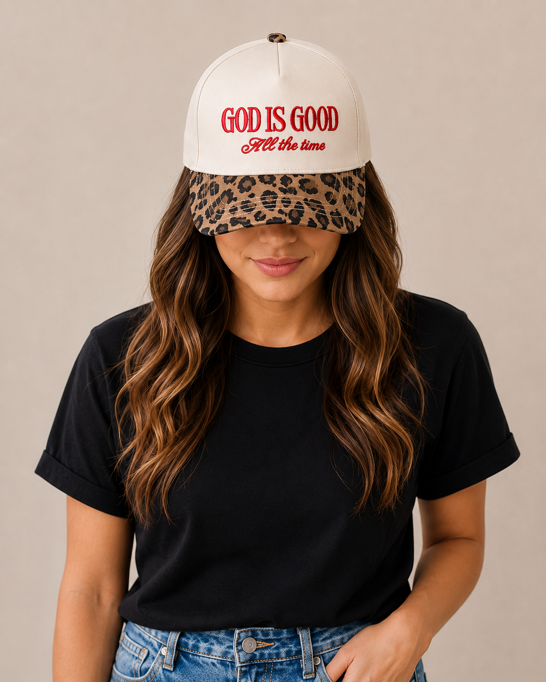 Person wearing a white cap with 'God is Good All the Time' text and a leopard print visor, black t-shirt, and blue jeans on a beige background.