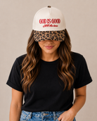 Person wearing a white cap with 'God is Good All the Time' text and a leopard print visor, black t-shirt, and blue jeans on a beige background.