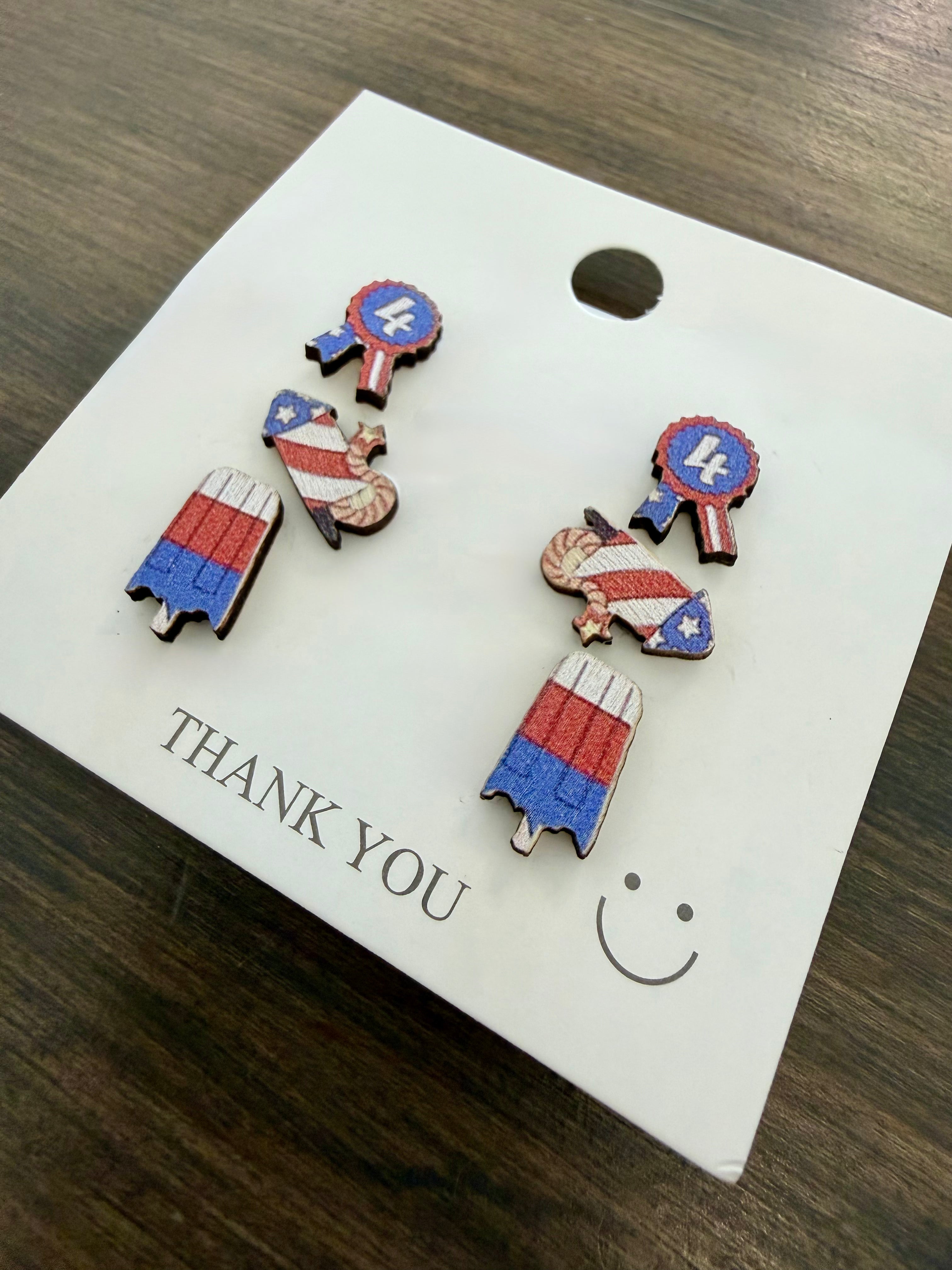 Set of 4th of July themed earrings on a 'THANK YOU' card with a smiley face on a wooden surface