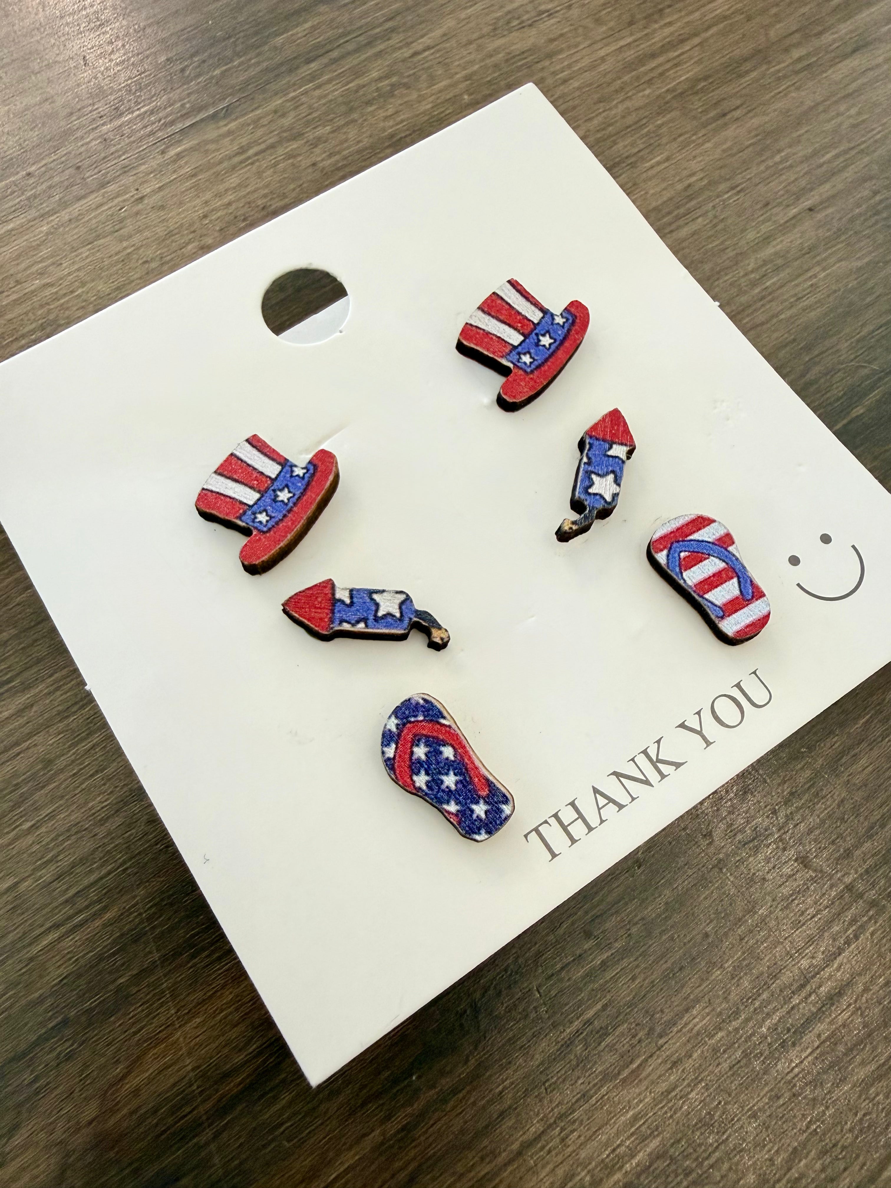 Set of 4th of July themed earrings on a 'THANK YOU' card with a smiley face on a wooden surface