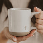 White mug with a cross symbol held by two hands against a neutral background