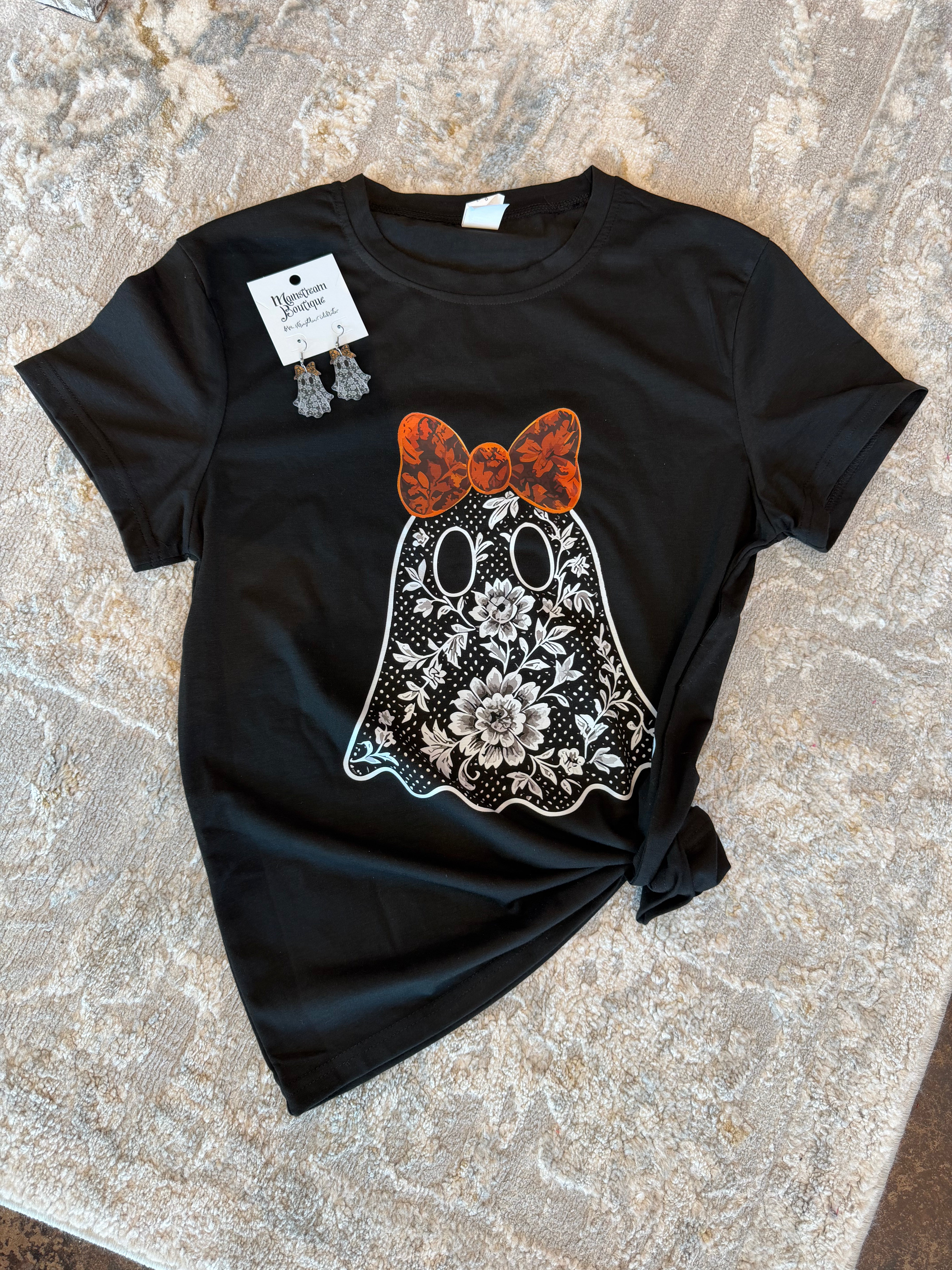 A black short-sleeve t-shirt with a ghost graphic featuring a floral Day of the Dead design and an orange bow, laid flat on a textured stone surface with a brand tag attached.