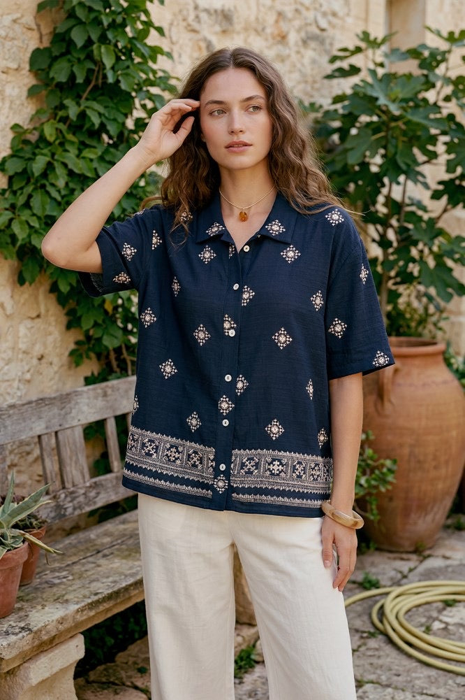 A woman stands outdoors in a rustic garden courtyard wearing a navy blue short-sleeve button-up shirt with white and beige bohemian-style geometric patterns, paired with cream-colored pants.