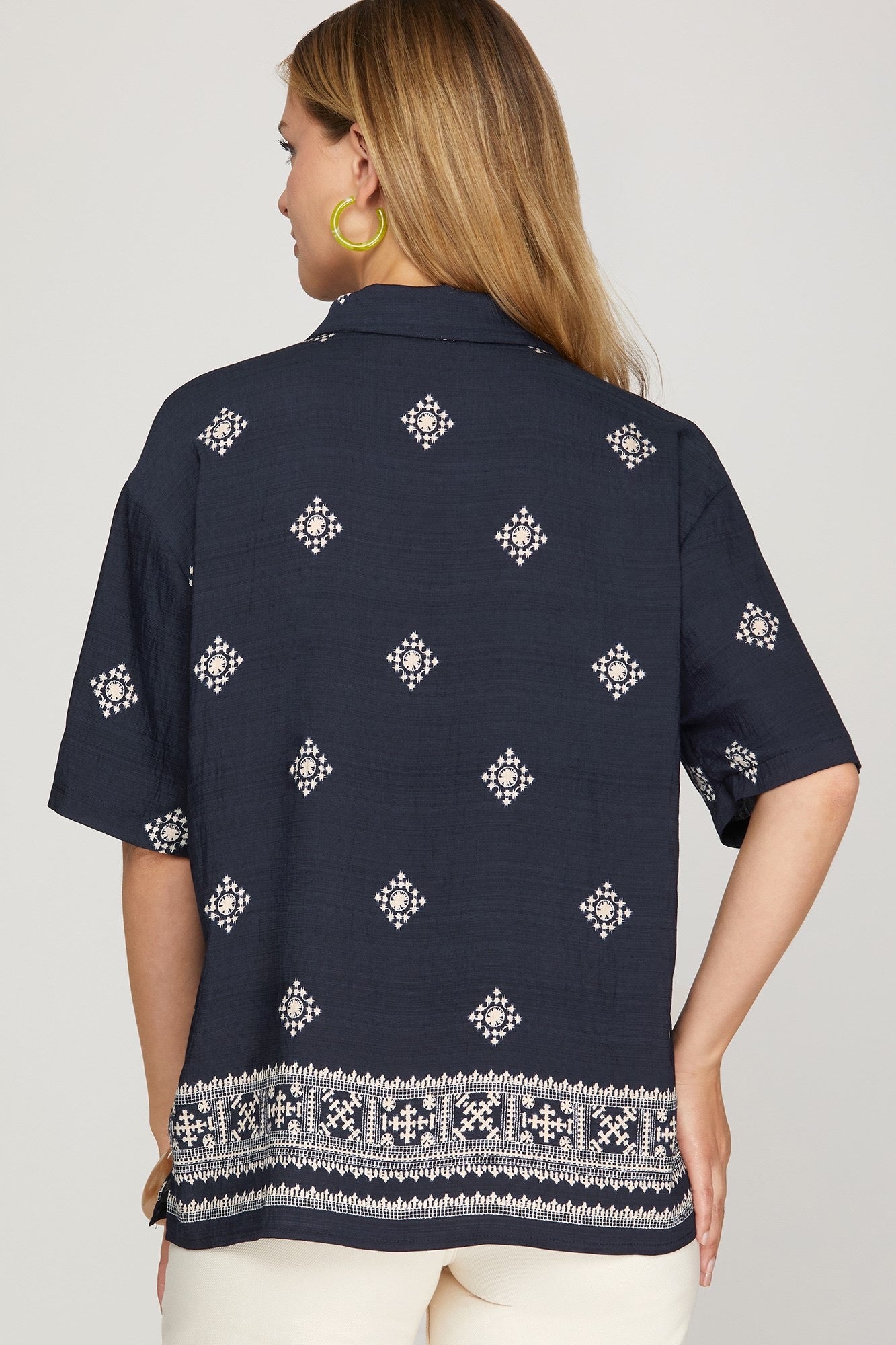 A woman shown from the back wearing a navy blue short-sleeve button-up shirt with white geometric patterns and a decorative border at the hem, paired with off-white pants against a light gray studio background.