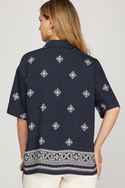 A woman shown from the back wearing a navy blue short-sleeve button-up shirt with white geometric patterns and a decorative border at the hem, paired with off-white pants against a light gray studio background.