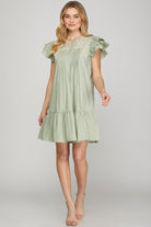 A woman modeling a light sage green tiered mini dress with ruffled cap sleeves and a lace-trimmed neckline, photographed against a neutral light gray studio background.