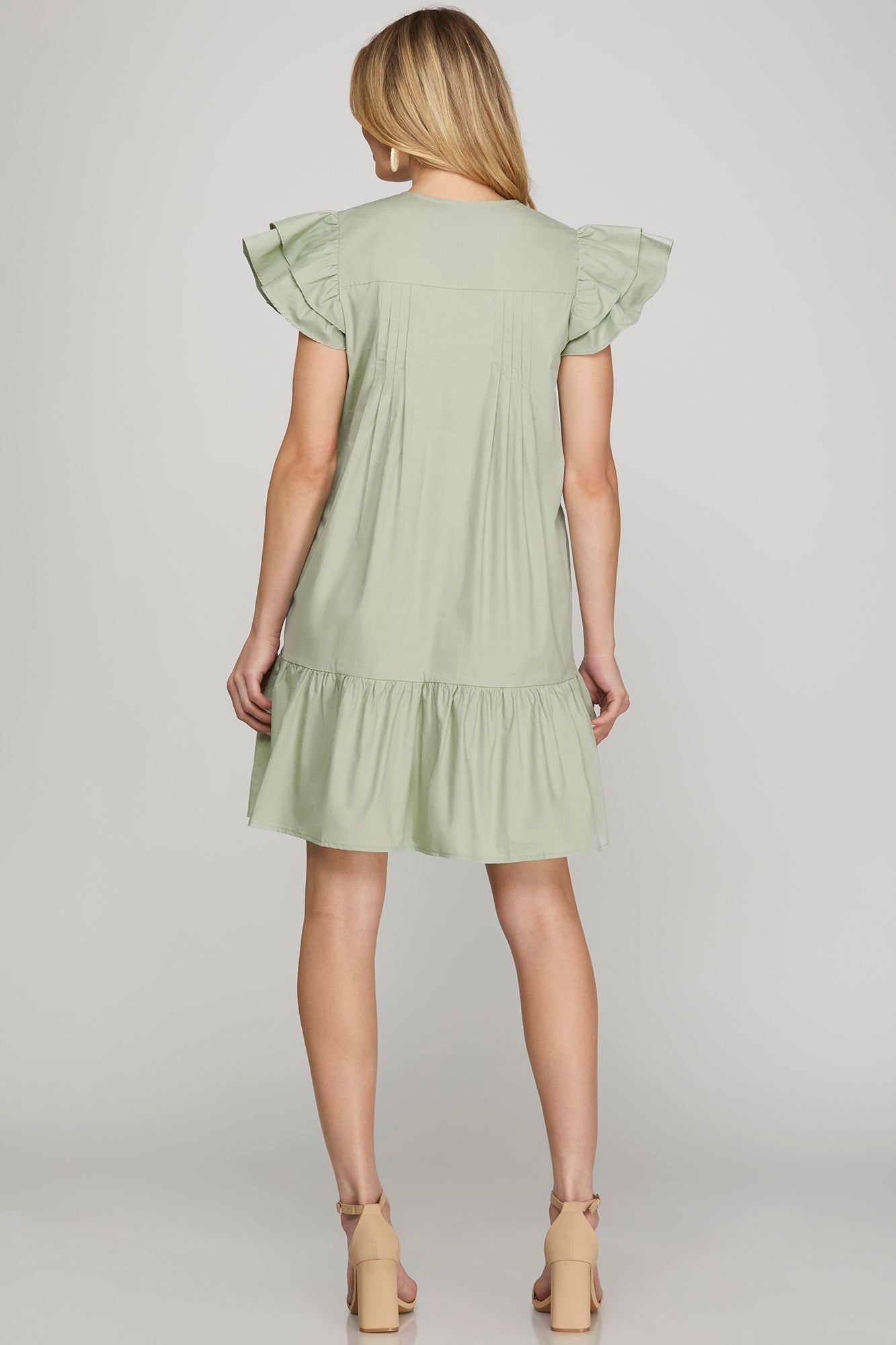 A woman shown from the back wearing a light sage green tiered tunic dress with ruffled cap sleeves, paired with beige high-heeled sandals, against a neutral gray studio background.
