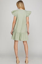 A woman shown from the back wearing a light sage green tiered tunic dress with ruffled cap sleeves, paired with beige high-heeled sandals, against a neutral gray studio background.