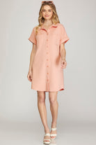 A woman modeling a short-sleeve button-front shirt dress in a soft peach-pink color, paired with white heeled sandals against a light gray studio background.