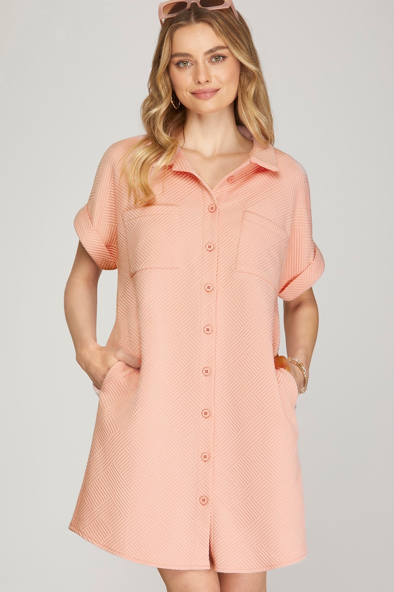 A woman modeling a rose-colored short-sleeve button-up shirt dress with a textured chevron pattern, set against a solid light gray background.