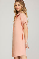 A woman wearing a short-sleeve, collared knit dress in a soft peach-pink color, photographed against a light gray studio background.