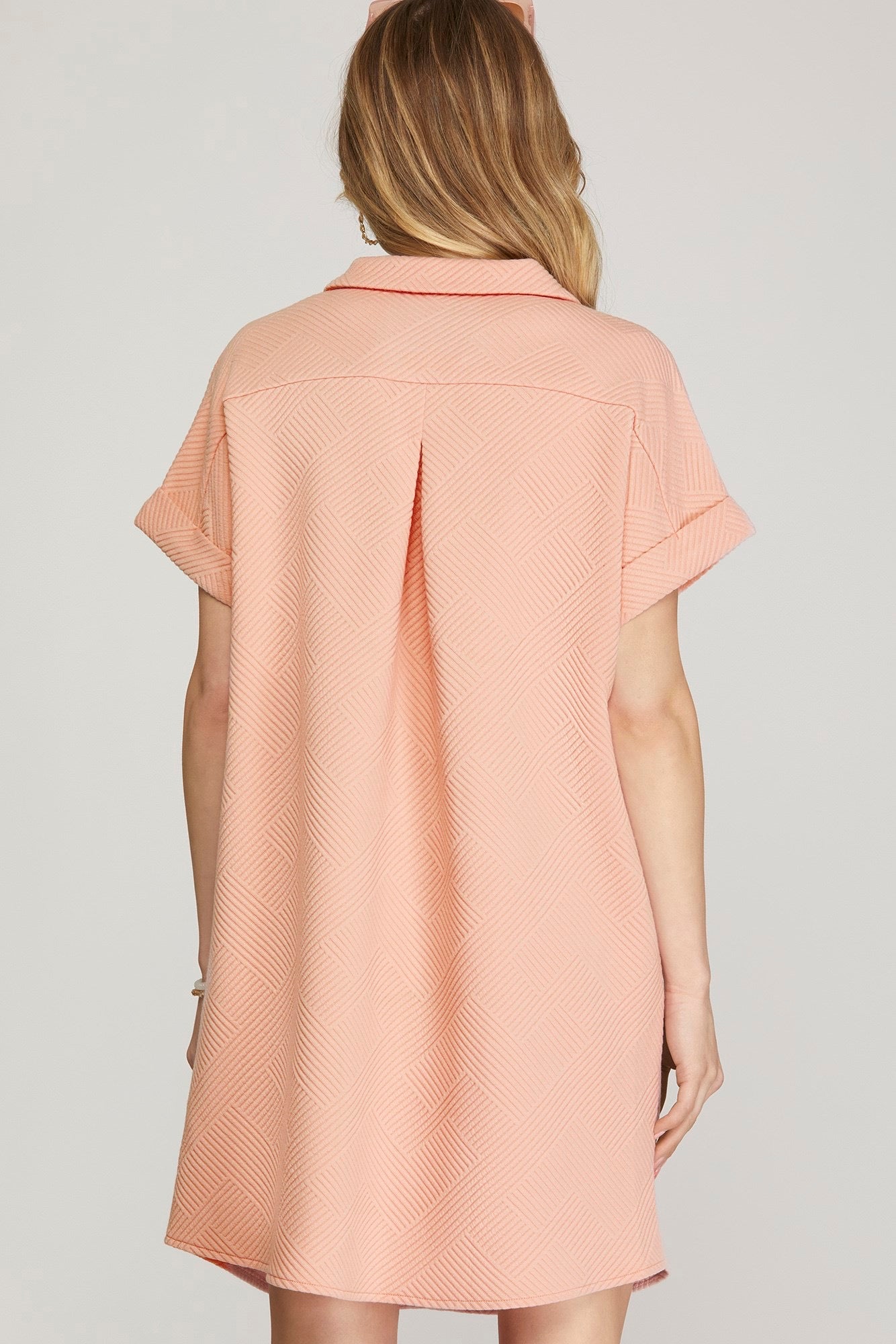 A woman shown from the back wearing a short-sleeve collared shirt dress in a light peach-pink color with a subtle textured pattern, set against a plain light gray studio background.