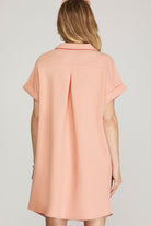 A woman shown from the back wearing a short-sleeve collared shirt dress in a light peach-pink color with a subtle textured pattern, set against a plain light gray studio background.