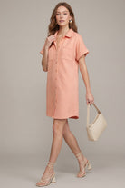 A woman models a short-sleeve peach-colored button-up shirt dress, styled with beige lace-up heeled sandals and a cream-colored shoulder bag, against a solid light gray studio background.