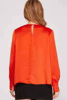 A woman viewed from the back wearing a bright orange satin blouse with a keyhole back detail and black skirt, against a light gray studio background.