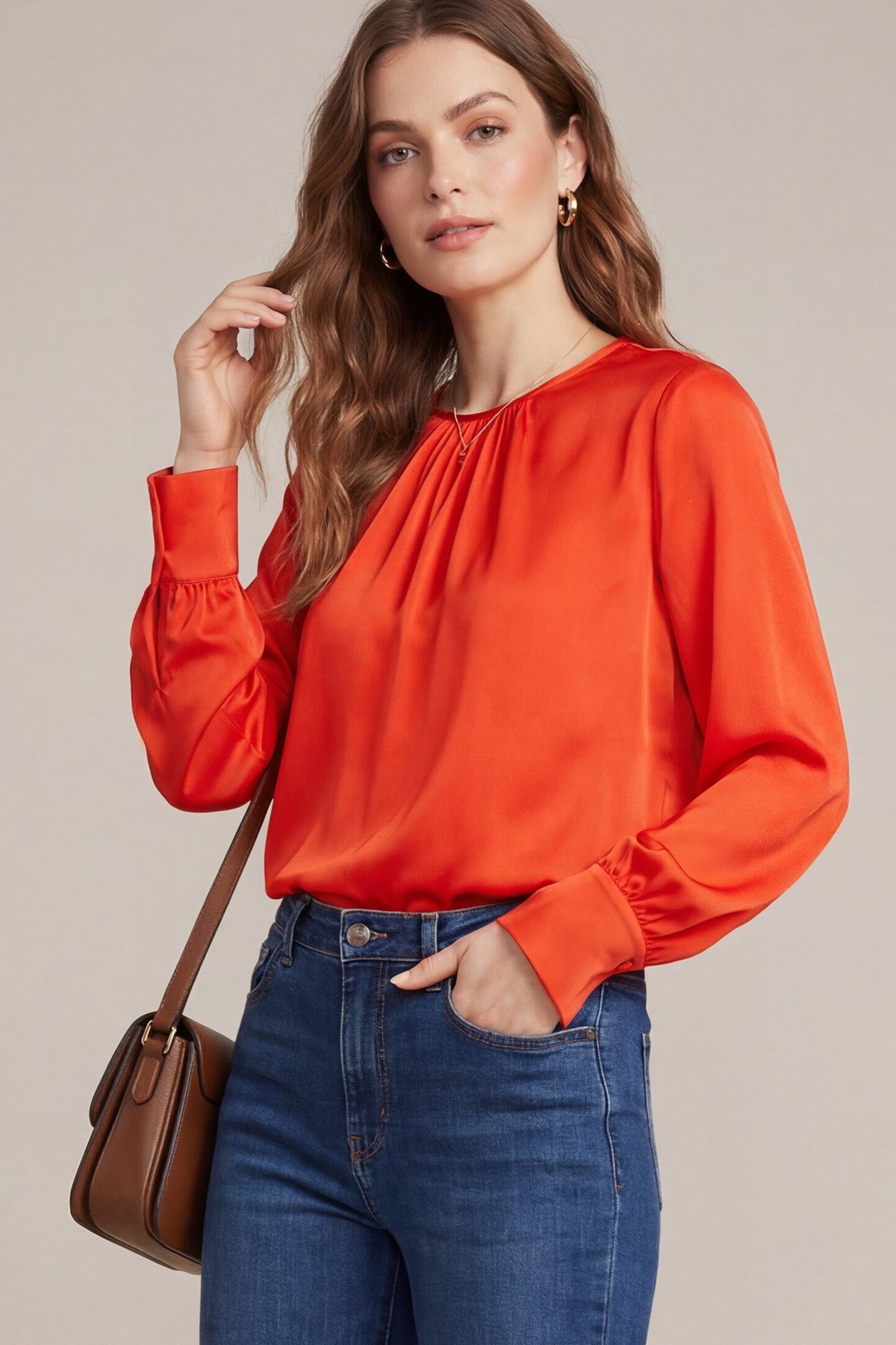 A woman wearing a bright orange long-sleeve blouse with a gathered neckline and elastic cuffs, paired with blue jeans and a brown shoulder bag, against a solid beige background.