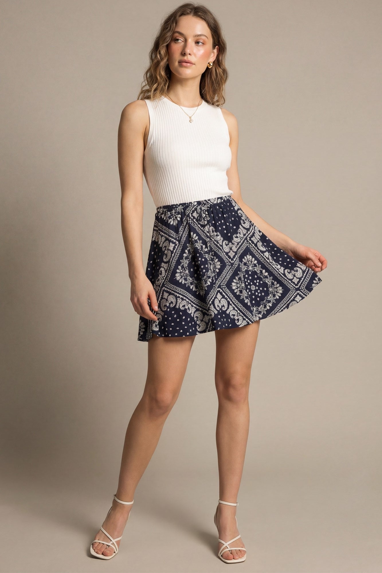 A woman modeling a sleeveless white ribbed knit top and a navy blue mini skirt with a white bandana print, standing against a solid beige studio background.