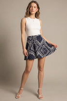 A woman modeling a sleeveless white ribbed knit top and a navy blue mini skirt with a white bandana print, standing against a solid beige studio background.