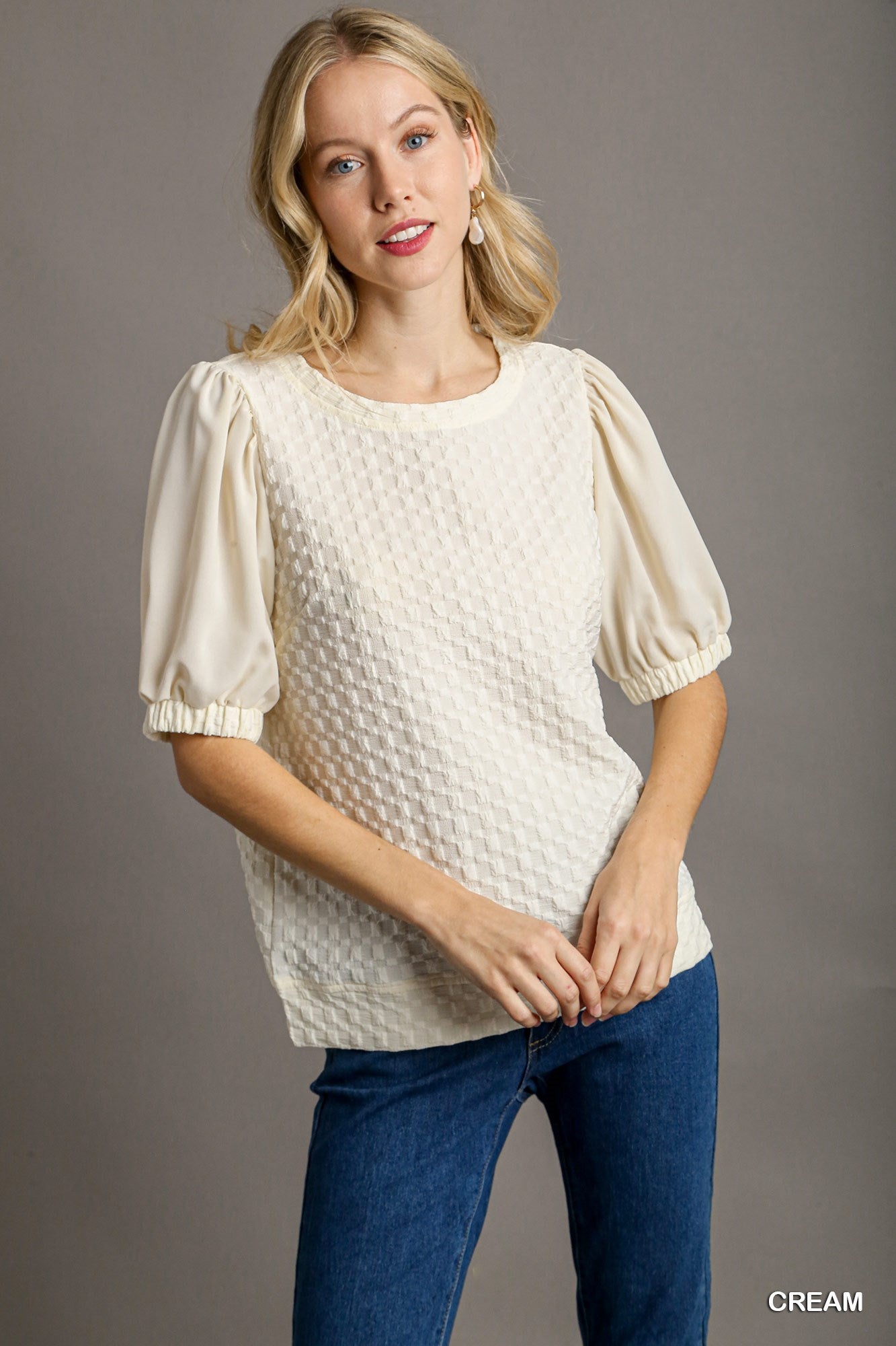 Woman wearing a cream textured blouse and blue jeans against a gray background