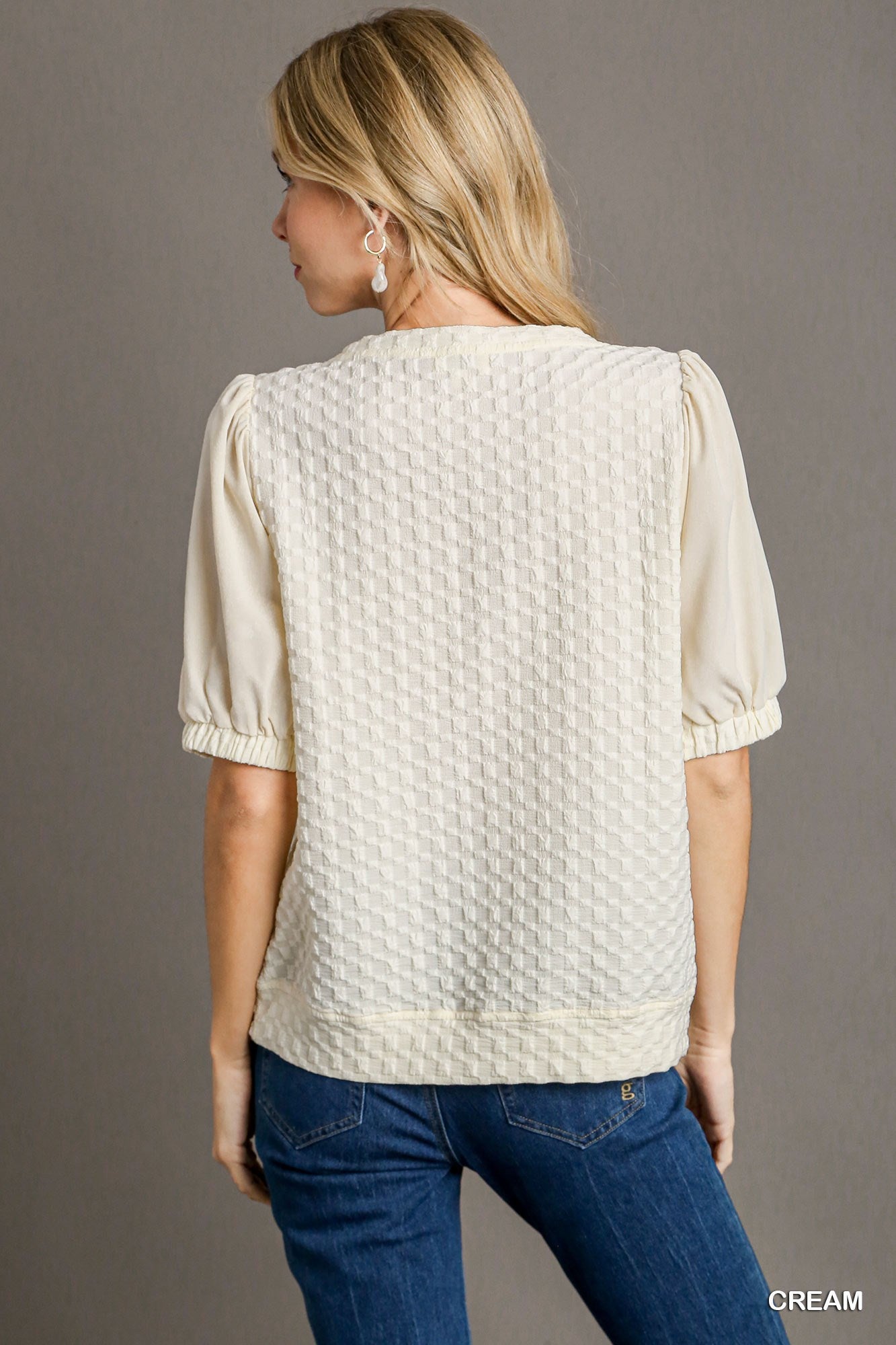 Woman wearing a cream textured blouse and blue jeans on a gray background