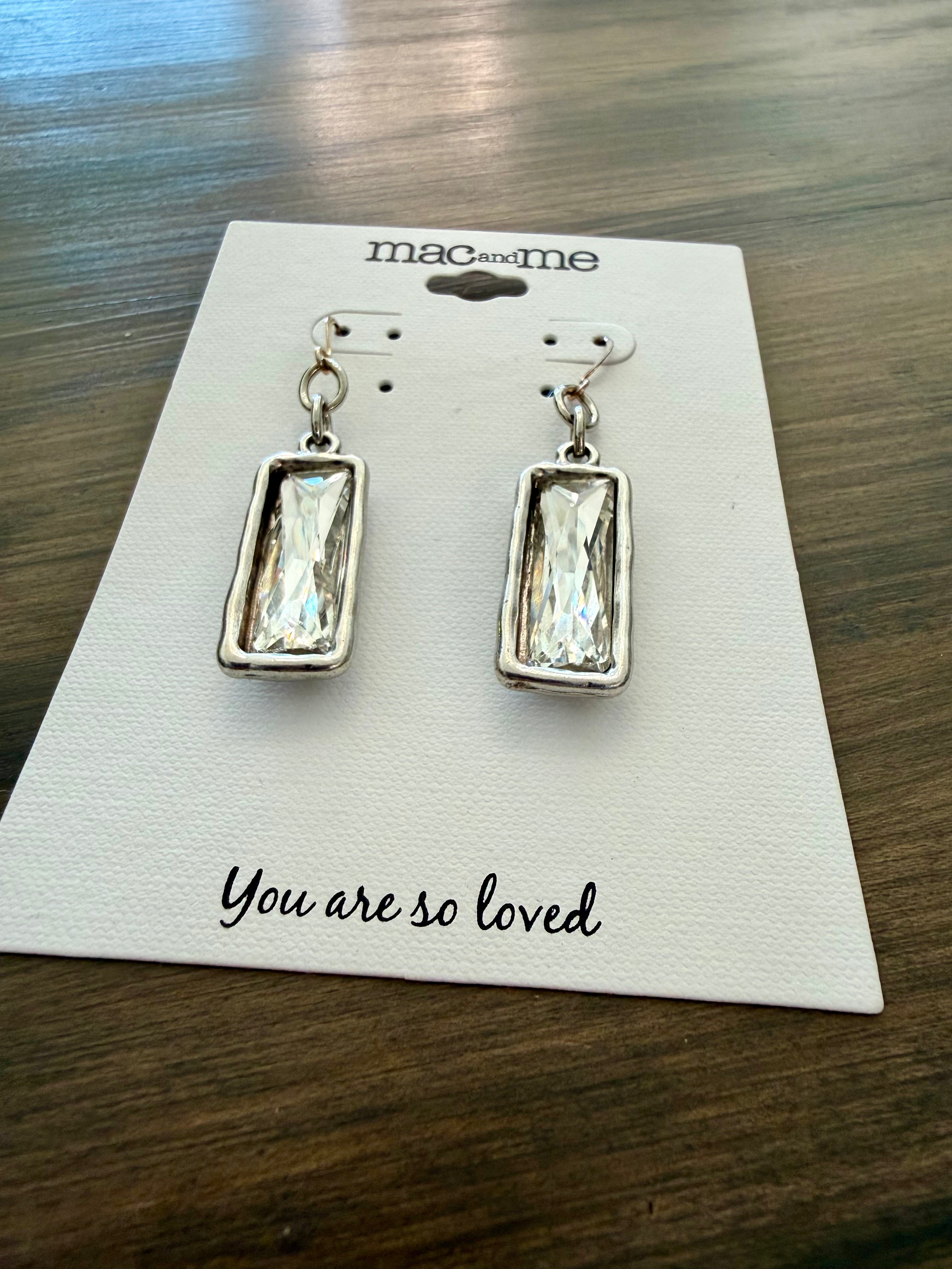 Silver earrings on a 'mac and me' branded card with 'You are so loved' text on a wooden surface.