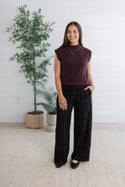A woman modeling a dark plum sleeveless knit top and black plaid wide-leg pants, standing in a bright room with a white shiplap wall, potted plants, and a light beige rug.