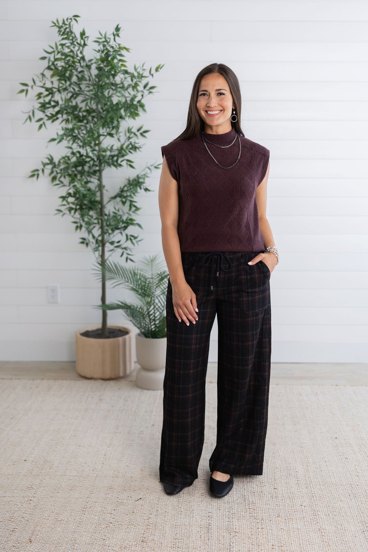 A woman modeling a dark plum sleeveless knit top and black plaid wide-leg pants, standing in a bright room with a white shiplap wall, potted plants, and a light beige rug.