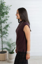 A woman shown in side profile wearing a sleeveless, ribbed knit top in a deep plum-burgundy color, paired with a dark plaid skirt. She is standing in a bright room with a white paneled wall and potted plants in the background.