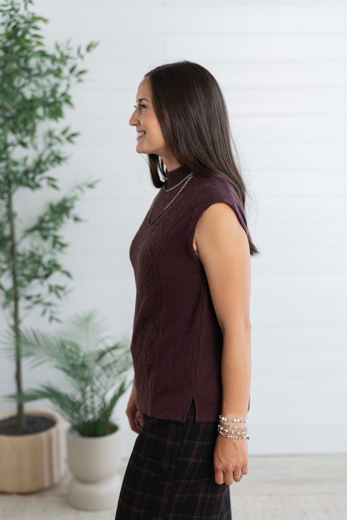 A woman shown in side profile wearing a sleeveless, ribbed knit top in a deep plum-burgundy color, paired with a dark plaid skirt. She is standing in a bright room with a white paneled wall and potted plants in the background.