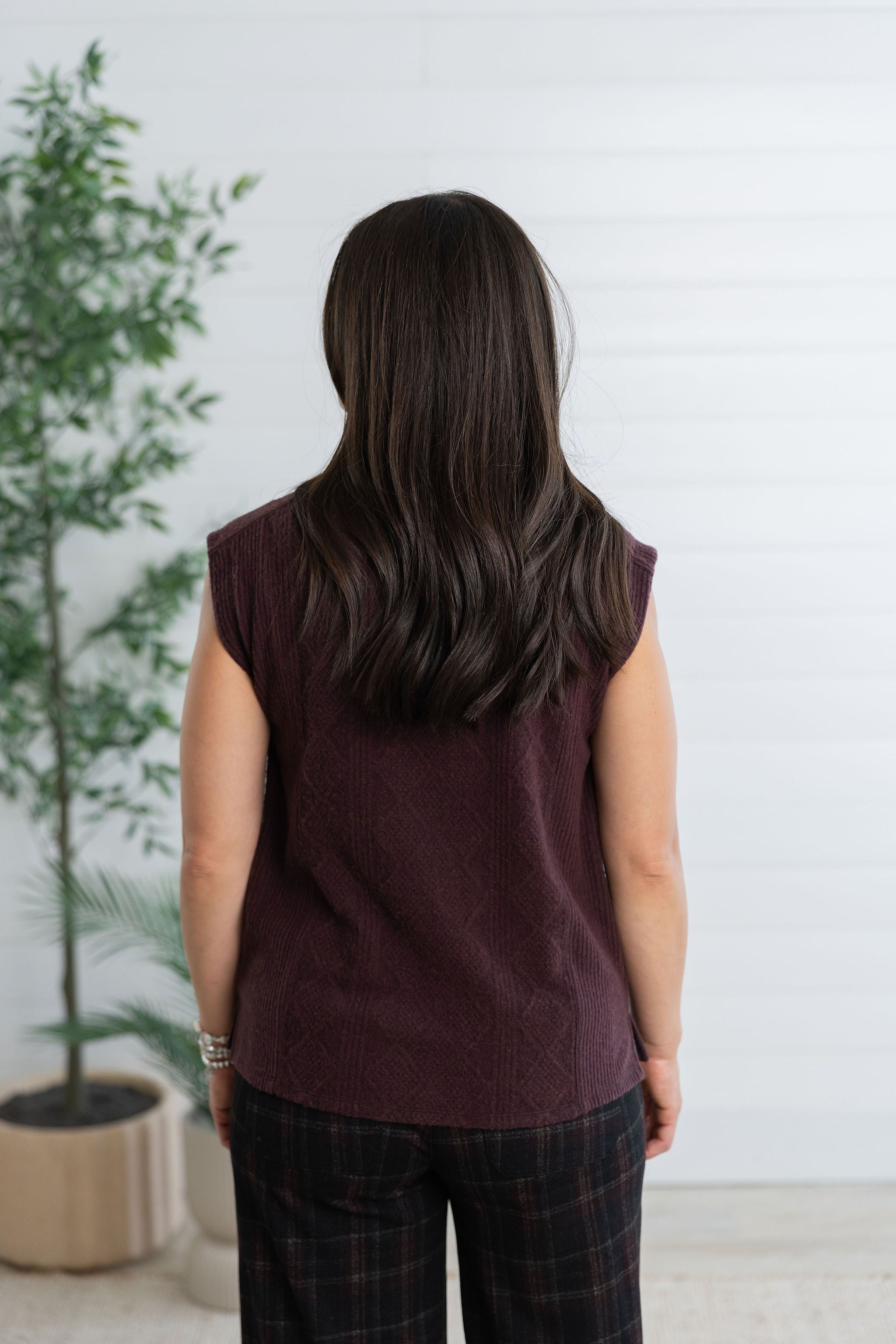 A woman viewed from behind wearing a sleeveless burgundy knit top and dark plaid pants, standing in front of a white paneled wall with a potted plant to the left.