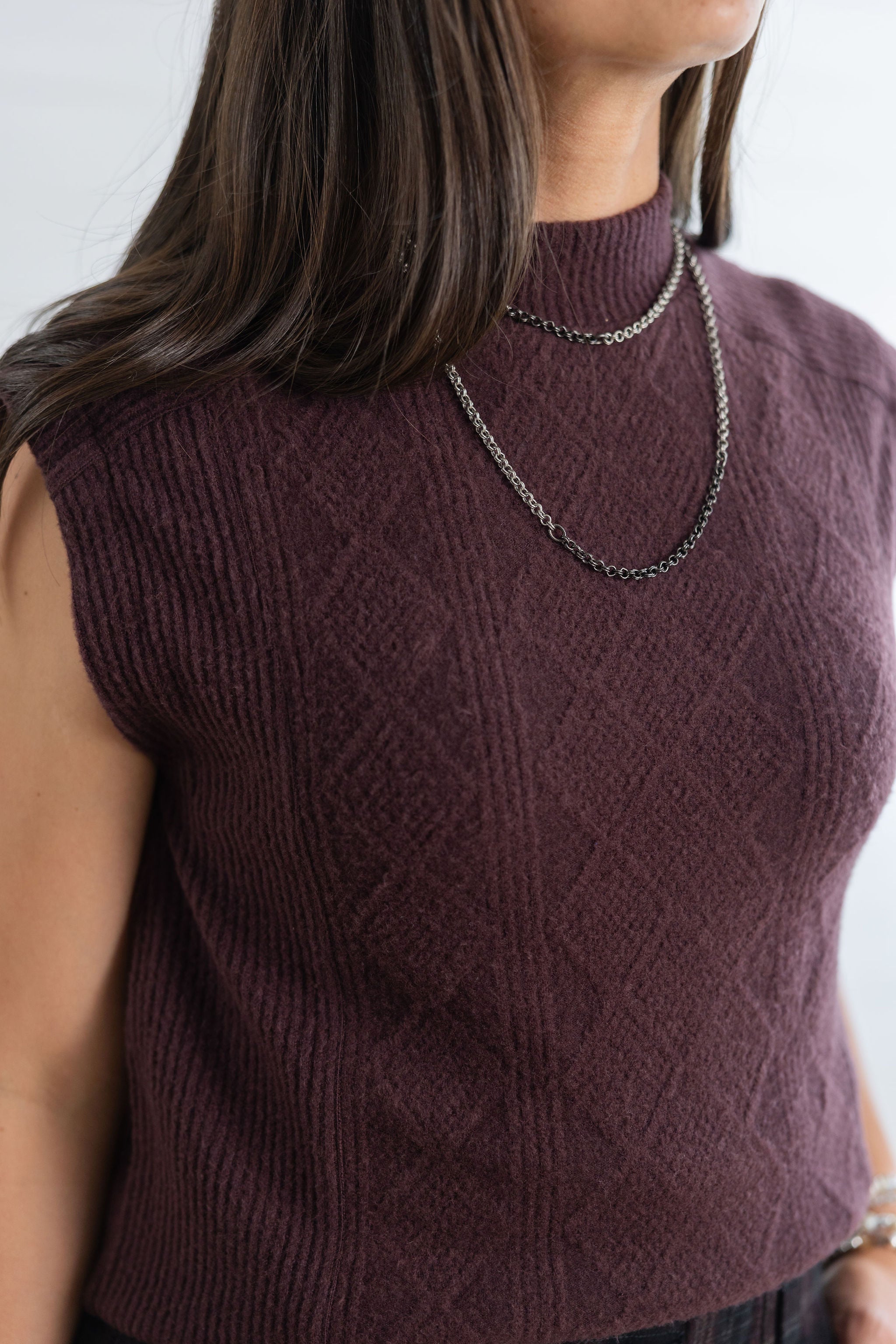 A close-up of a woman wearing a sleeveless ribbed knit mock neck top in a deep plum-burgundy color, styled with a silver chain necklace.