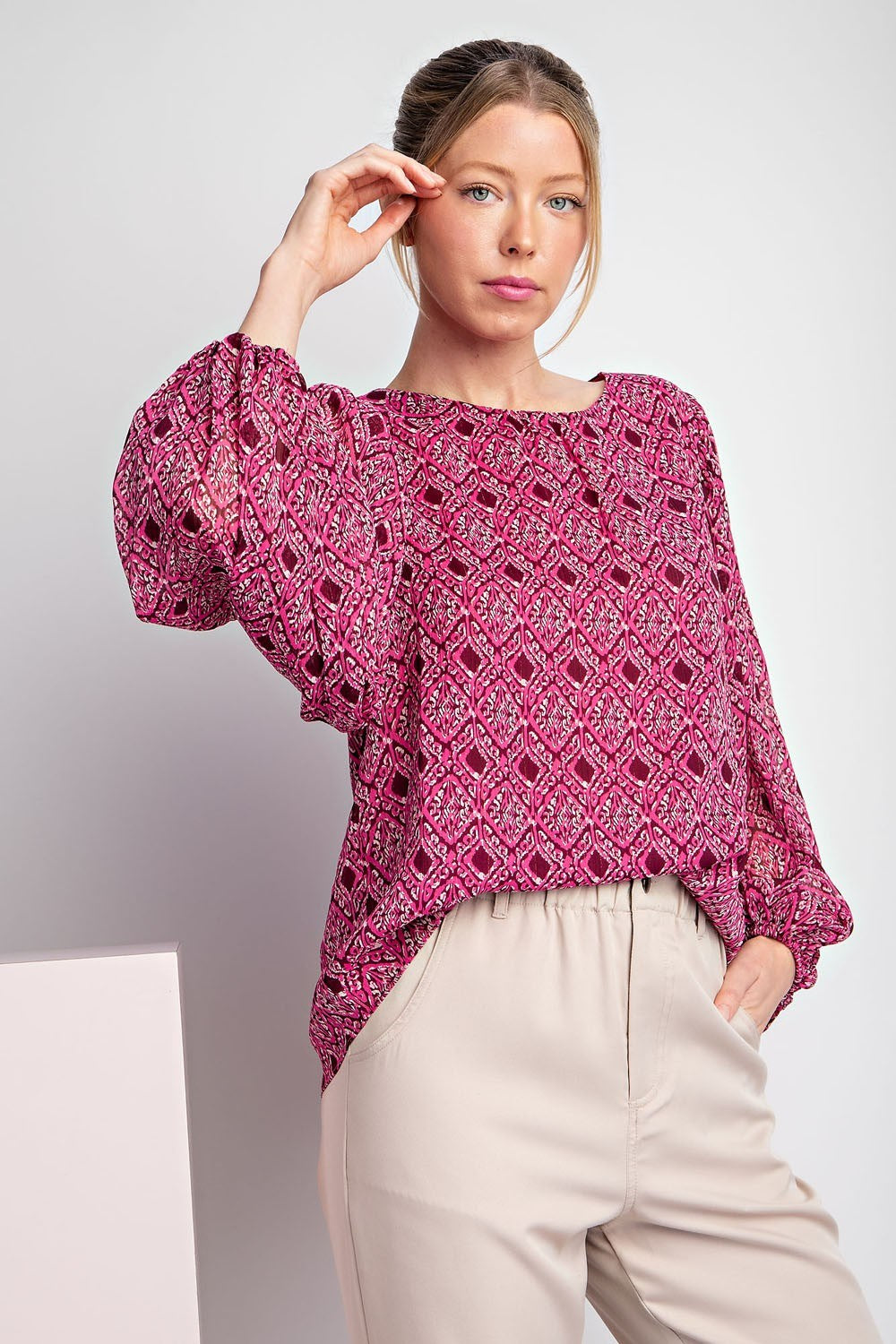 Woman wearing a pink patterned blouse and beige pants against a white background